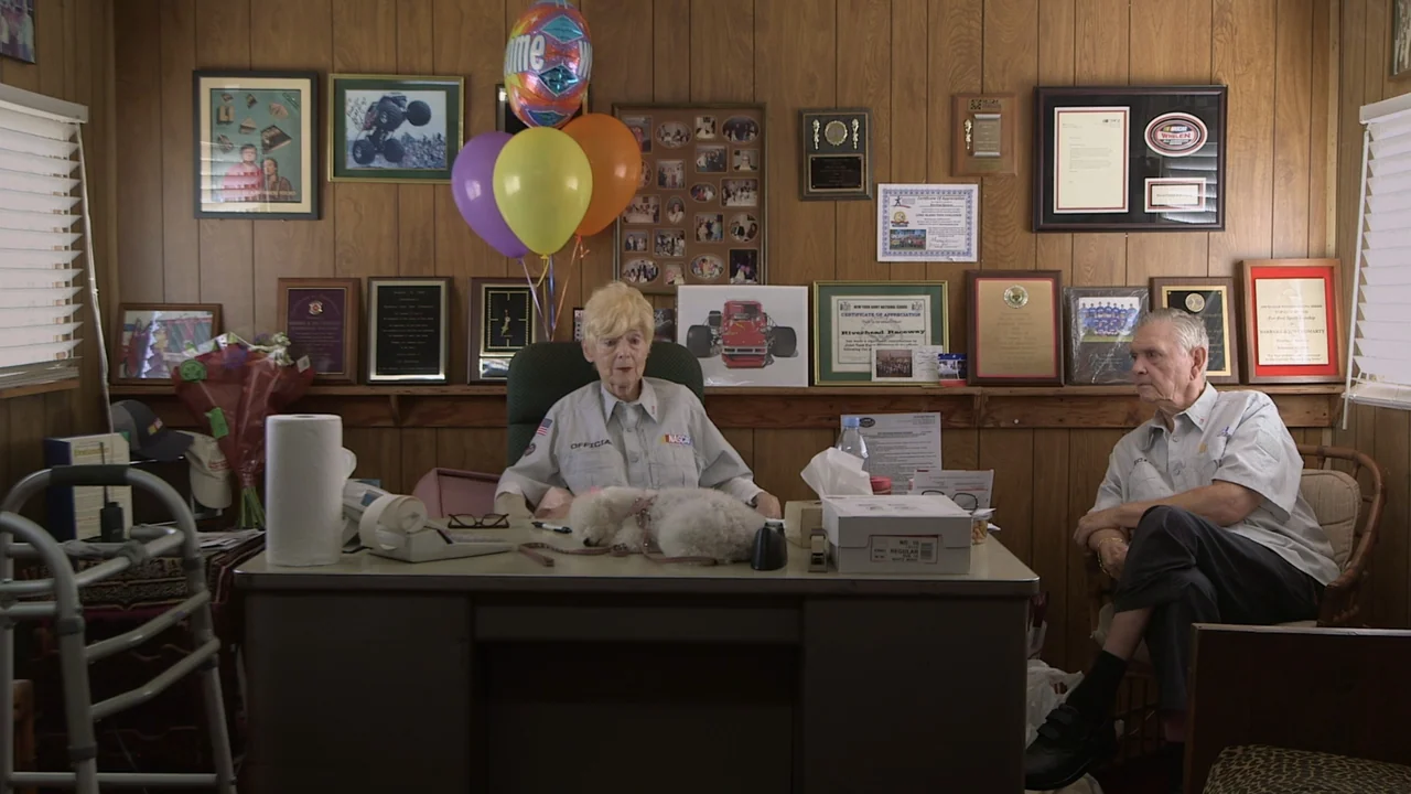 The Last Race (2022) movie still: medium — An office with wood-paneled walls decorated with framed certificates, trophies, and colored balloons. An…
