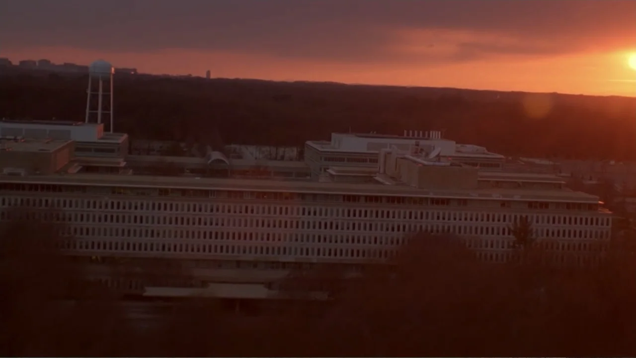 The Forever Prisoner (2021) movie still: establishing — A large, low-profile mid-century modern office complex sits beneath a setting sun. A water…