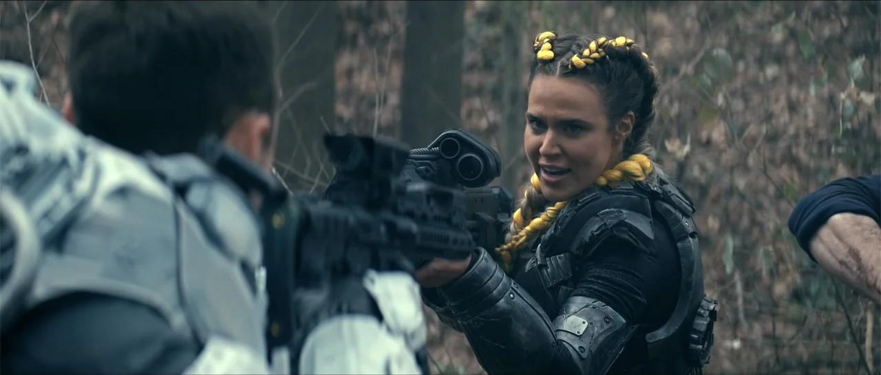 Cosmic Sin (2021) movie still: medium — A dense outdoor wooded area with blurred bare trees in the background. A woman in tactical armor aims a…