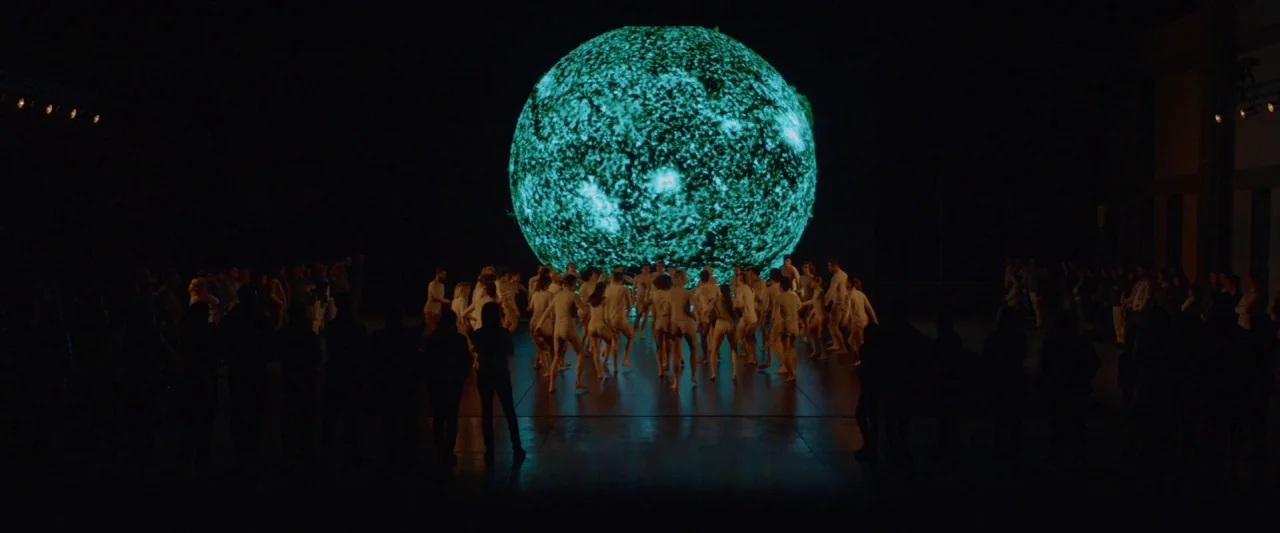 Ema (2019) movie still: wide — A large dark performance space features a glowing sphere displaying the sun's surface. Dancers in white garments are…