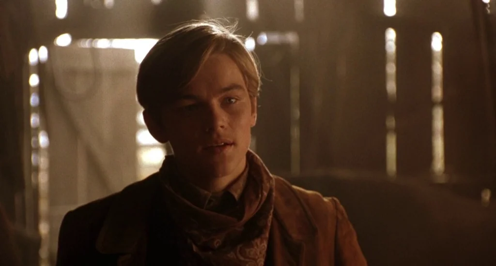 The Quick and the Dead (1995) movie still: medium — A dimly lit wooden structure with light streaming through vertical wall gaps. A young man stands…