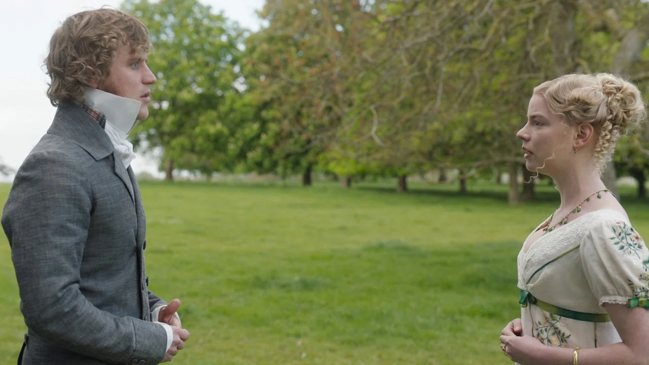 Emma. (2020) movie still: medium — Two individuals stand in a grassy open field surrounded by mature deciduous trees. A man in a grey suit and a…