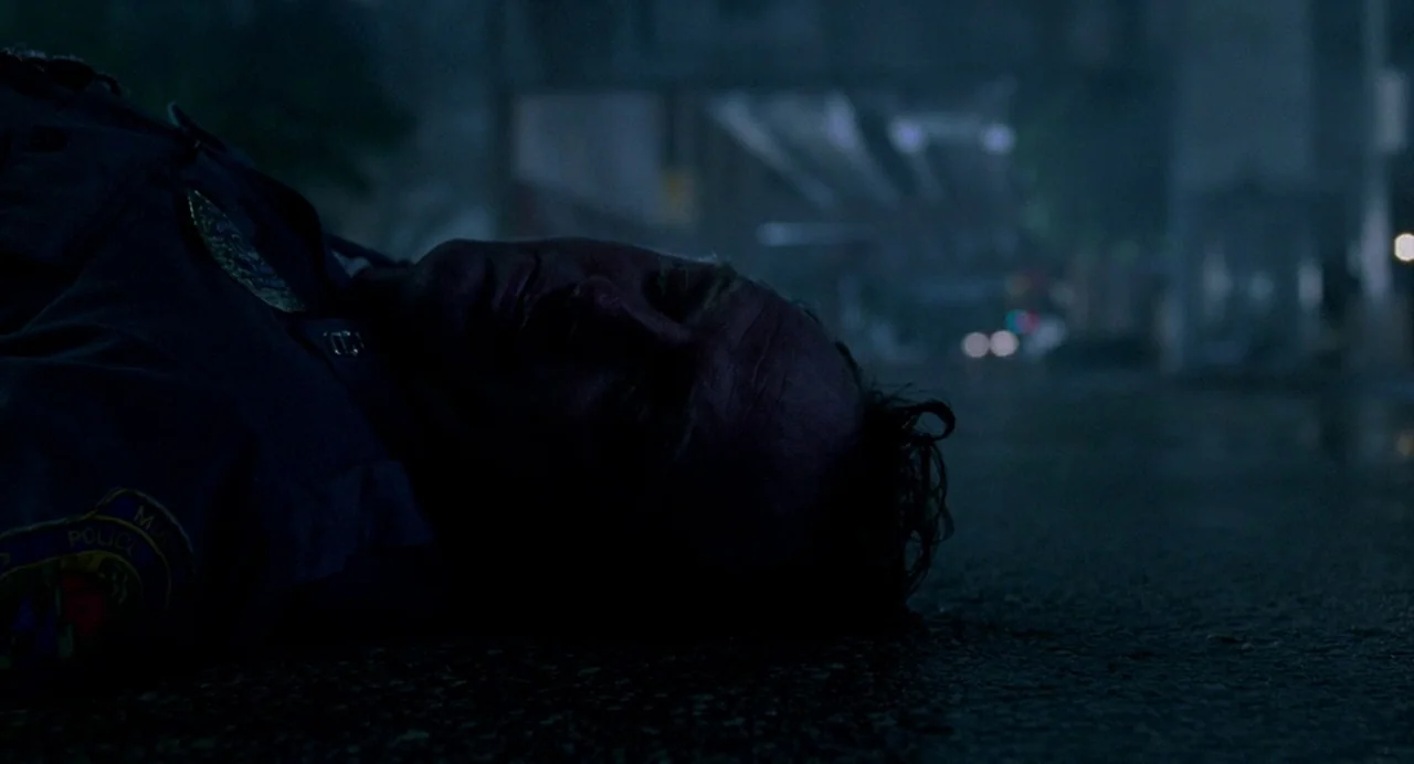 Bad Boys (1995) movie still: closeup — A man in a dark police uniform lies motionless on wet asphalt at night. Background elements suggest an urban…