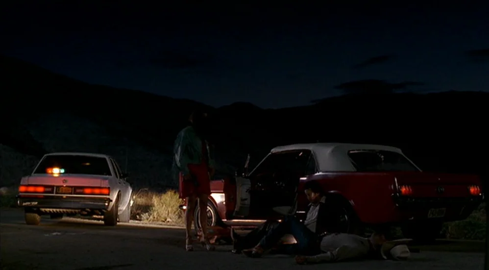 U-Turn (1997) movie still: wide — A desolate paved road at night features two vehicles stopped side by side. One person stands while two others are…