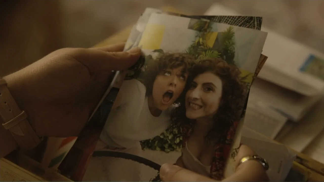 Lecciones para canallas (2022) movie still: closeup — A person holds a color photograph depicting two women in an indoor setting. The photograph…