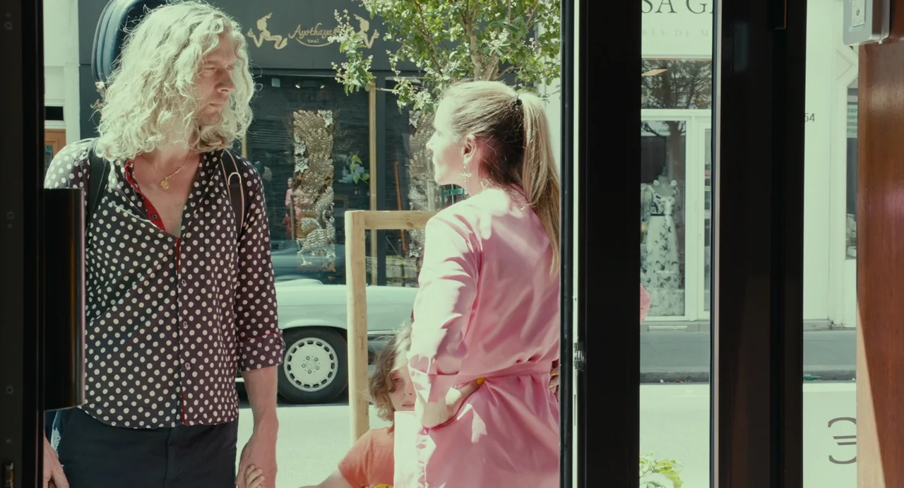 Last Summer (2023) movie still: medium — A man with long curly hair and a woman in a pink dress stand outside. They are positioned on a street in…