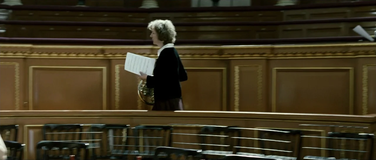 The Oxford Murders (2008) movie still: medium — An interior concert hall features ornate wood-paneled walls and rows of empty black chairs. A woman…
