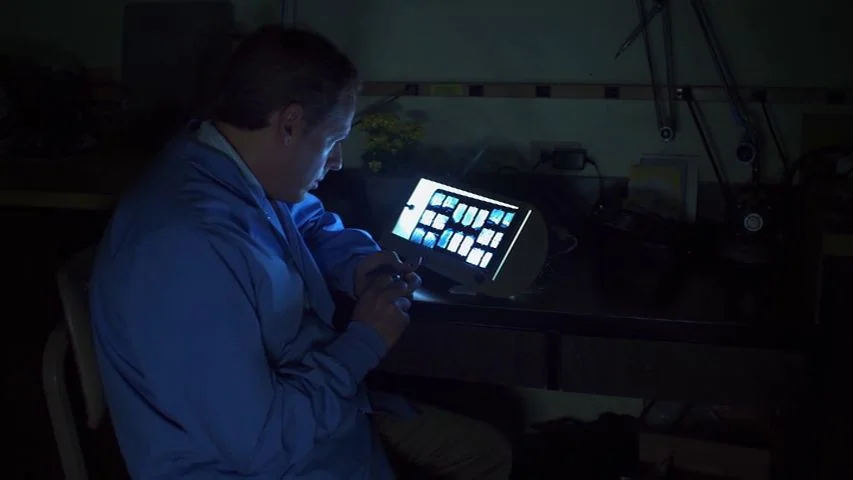 Touchy Feely (2013) movie still: medium — A dimly lit room contains a desk holding technical equipment and a light box. A man sits at the desk,…