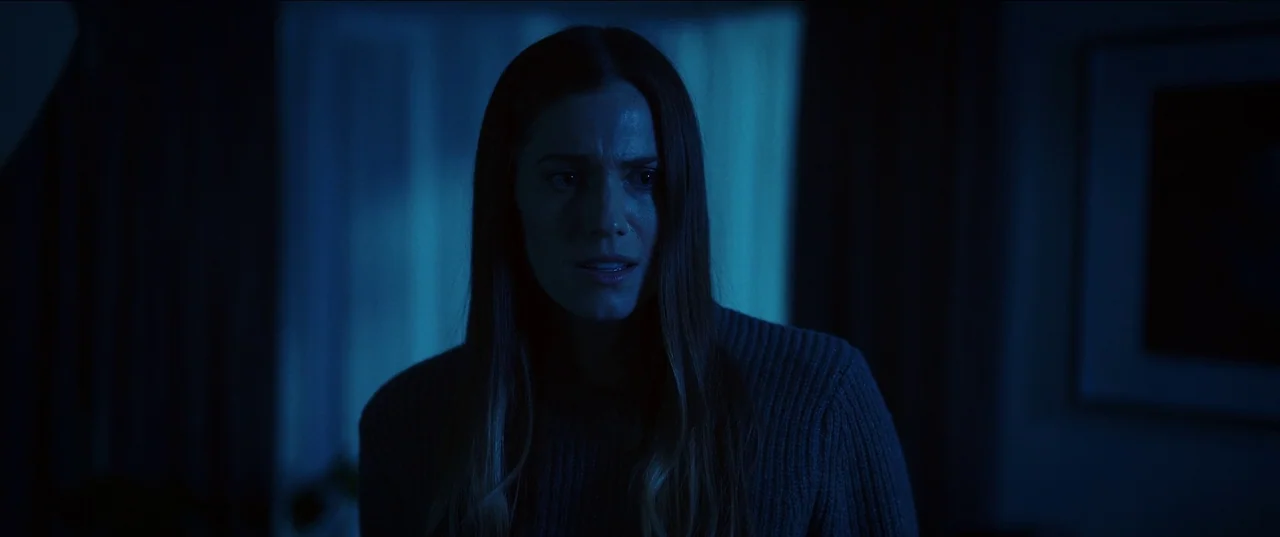 M3gan (2022) movie still: medium — A room at night is bathed in cold, blue-tinted light from an unseen window. A woman looks off-screen with a…