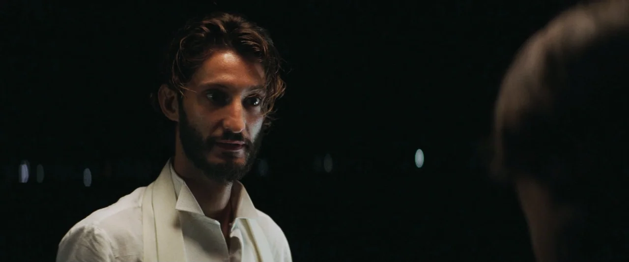 Masquerade (2022) movie still: medium — A dark outdoor environment with distant out-of-focus light points visible in the background. A man with dark…