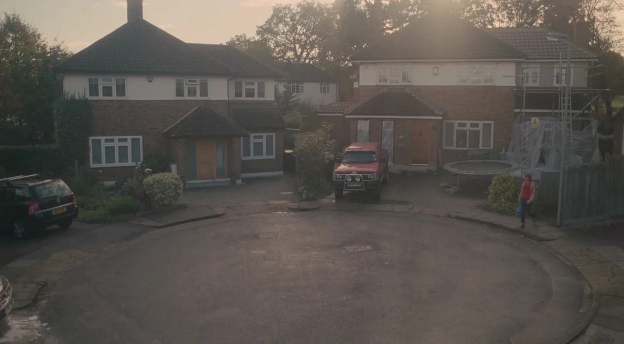 Broken (2012) movie still: wide — A suburban cul-de-sac features two brick houses with driveways at dusk. A woman walks along the pavement near a…