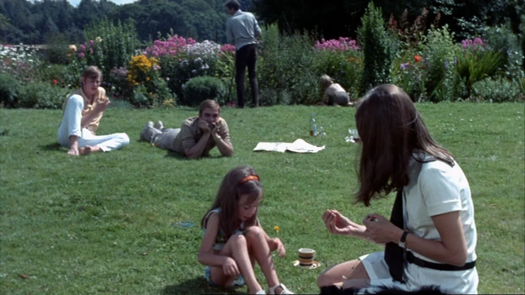 Accident (1967) movie still: wide — A group of people relaxes on a sunny lawn during a summer afternoon. The garden features flower beds and trees…