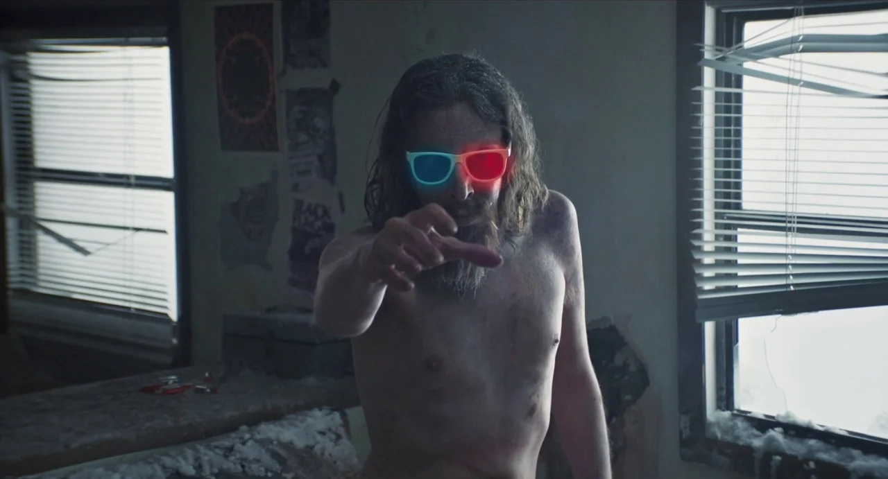 Relaxer (2018) movie still: medium — A dilapidated room features peeling paint and posters on the wall between two windows. A shirtless man with…