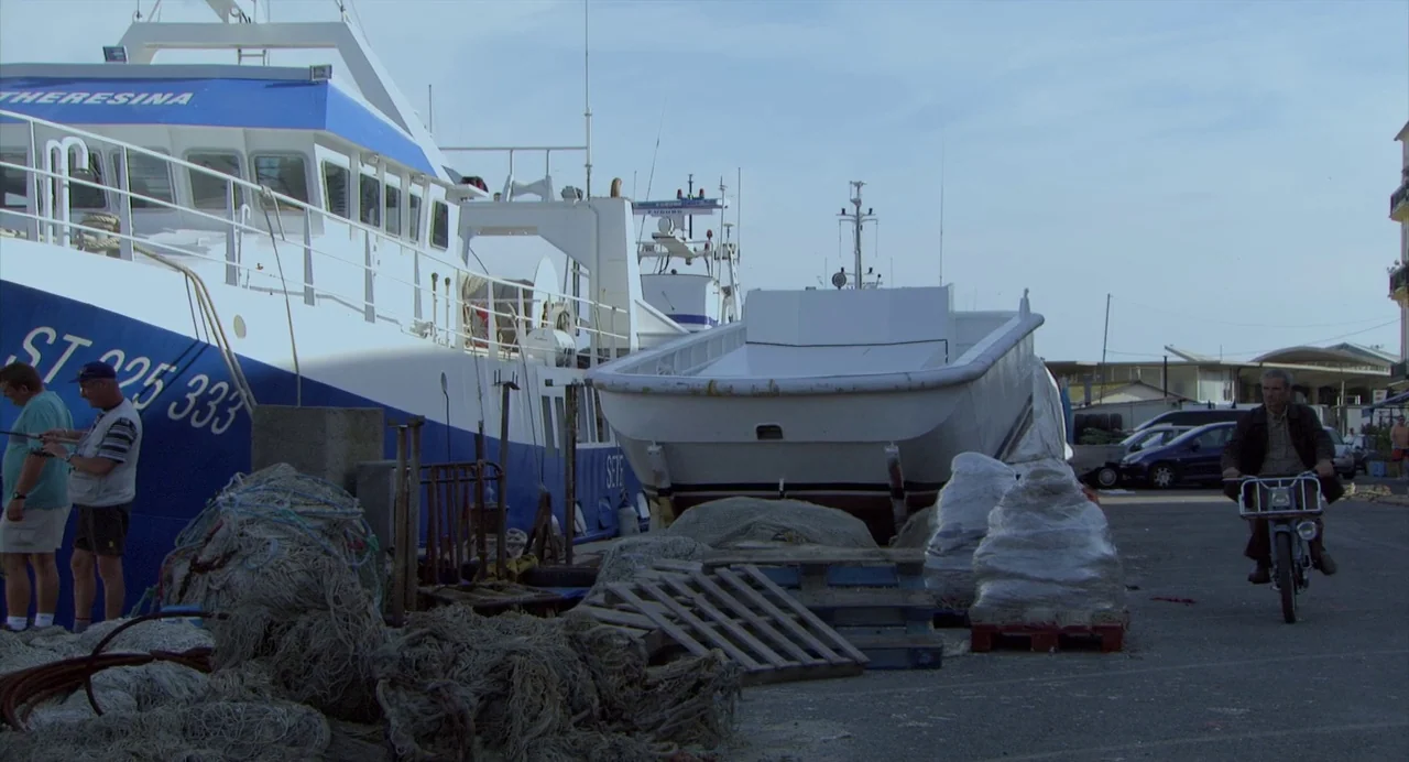 The Secret of the Grain (2007) movie still: wide — A fishing port dock holds large white vessels, fishing nets, and stacked wooden pallets. A man on…