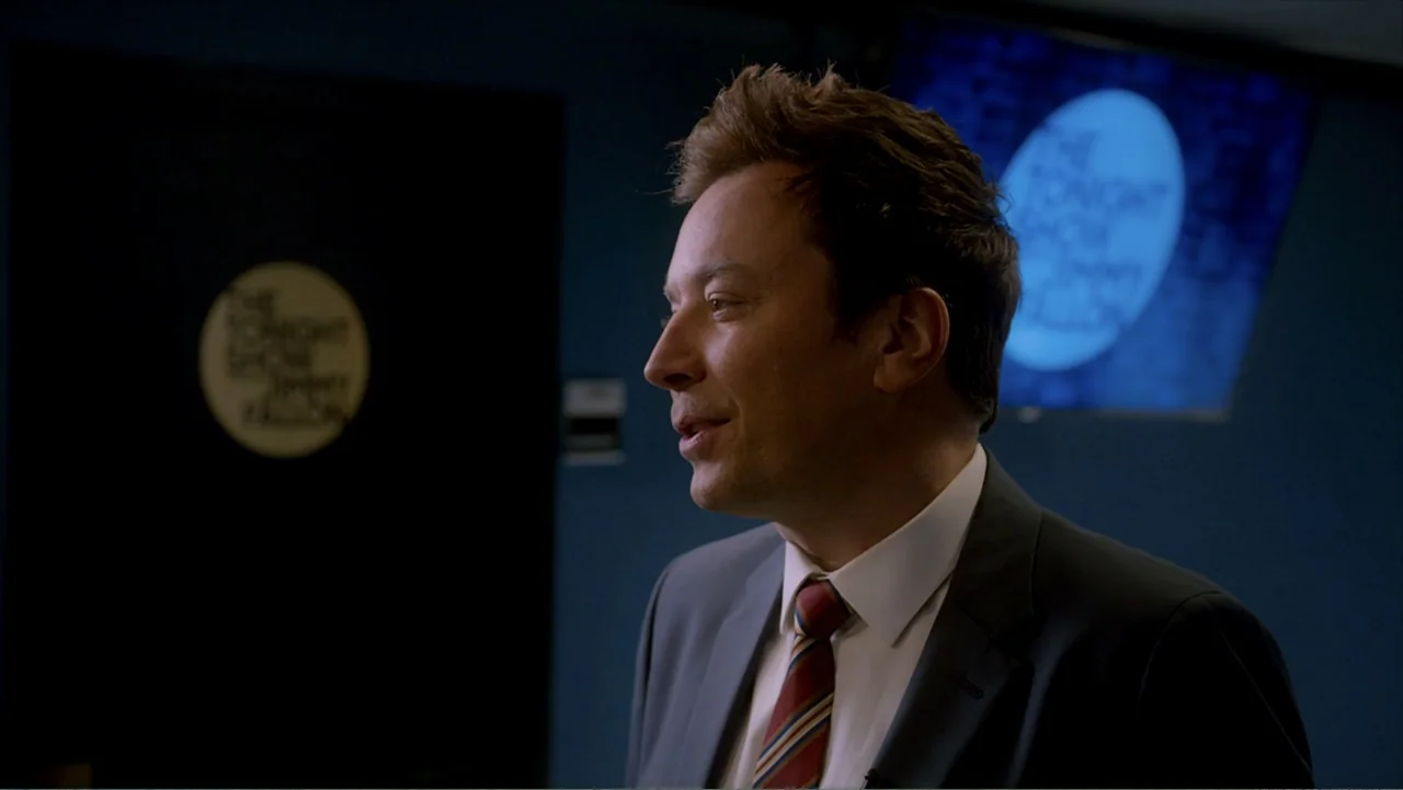 Jack in Time for Christmas (2024) movie still: medium — A dimly lit television studio features a dark blue wall with circular logo signage. A man in…