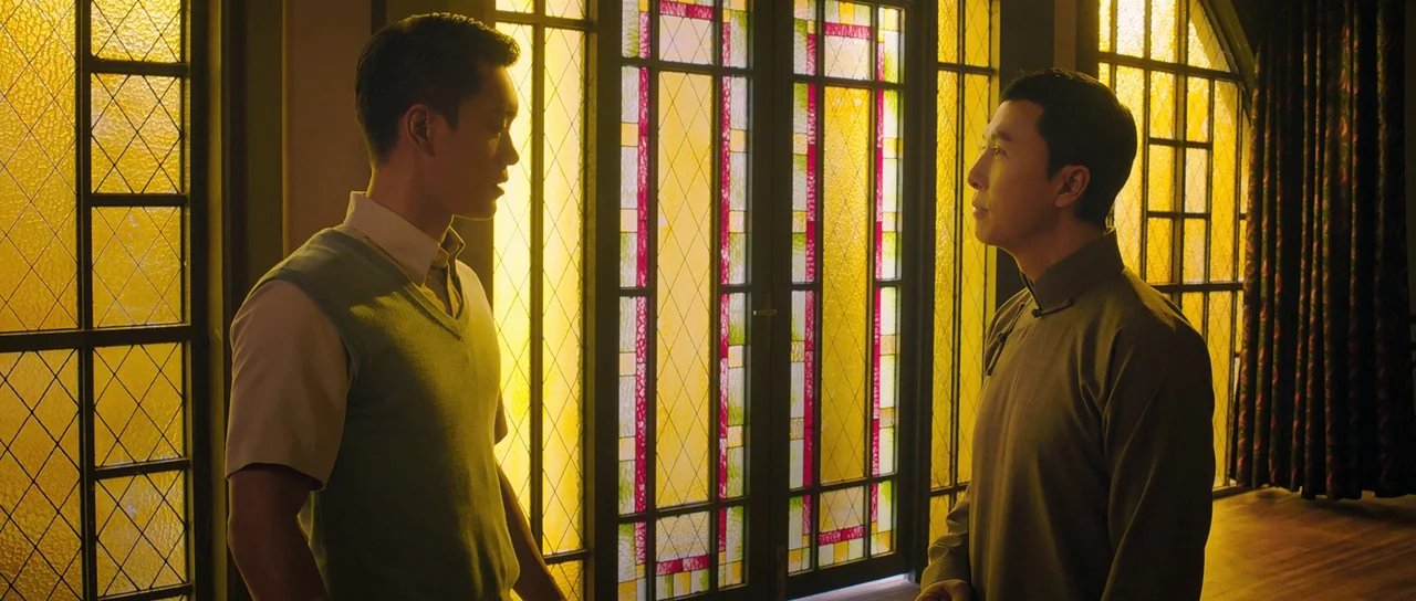 Ip Man 3 (2015) movie still: medium — An interior space features tall decorative stained glass windows with a geometric grid pattern. Two men stand…