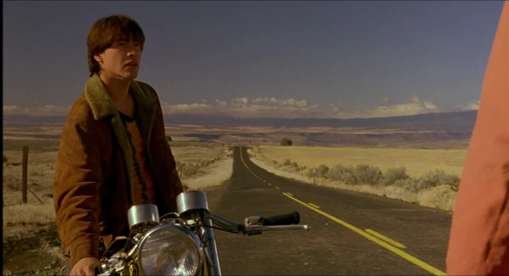 My Own Private Idaho (1991) movie still: medium — A desolate desert road stretches toward distant mountains under a clear, bright sky. A man sits on…