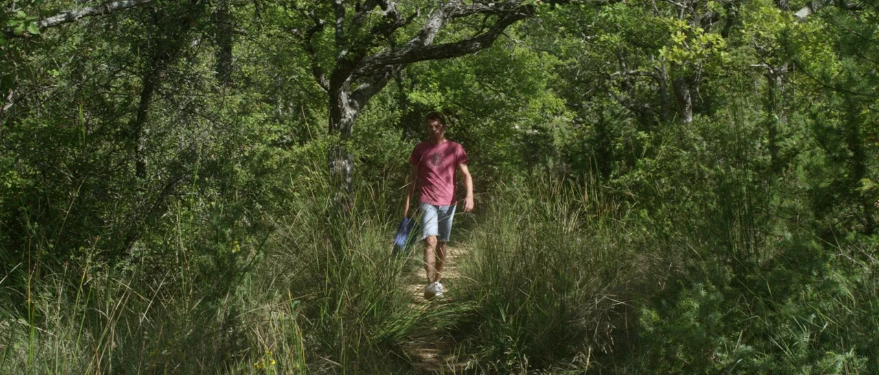 Stranger By The Lake (2013) movie still: fullbody — A narrow dirt trail cuts through a dense forest filled with tall wild grasses. A young man walks…