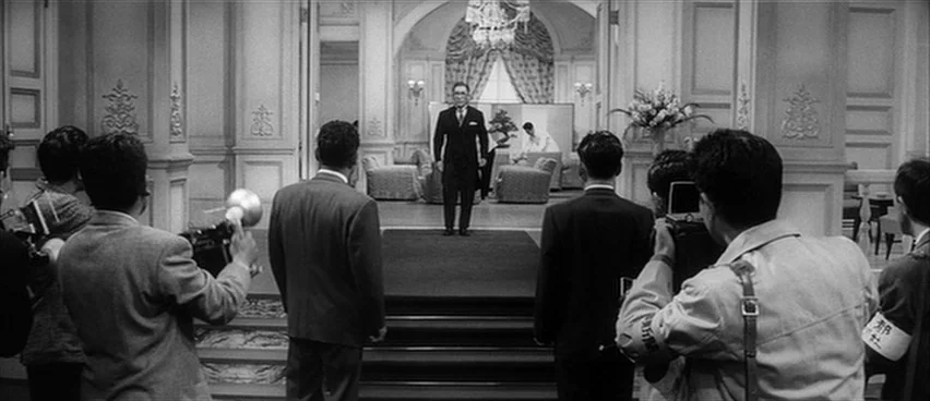 The Bad Sleep Well (1960) movie still: wide — A grand, formal space with white paneled walls and marble flooring leads to an archway. A man stands…