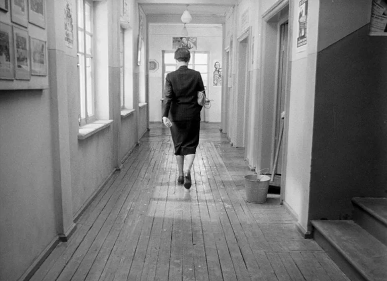 Wings (1966) movie still: fullbody — A long, narrow institutional hallway features wooden floorboards and windows lining one side wall. A lone woman…
