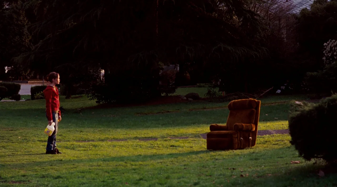 Juno (2007) movie still: wide — A teenage girl stands on a grassy lawn during golden hour. A single brown armchair sits on the grass a few feet away…