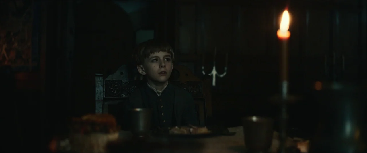 The King movie still: medium — A dark, wood-paneled medieval dining hall with minimal furniture and heavy shadows. A young boy sits alone at a table…