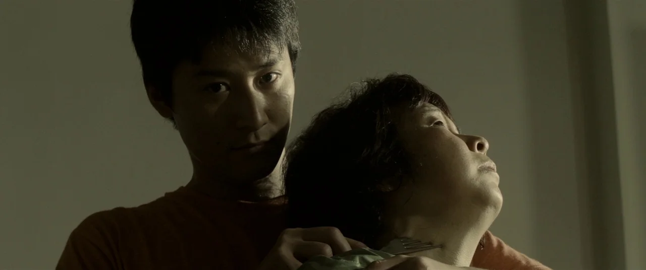 Creepy (2016) movie still: medium — A dimly lit room provides the backdrop for a confrontation between two individuals. The man stands behind the…
