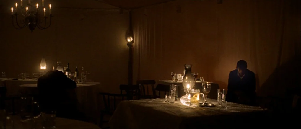 The Claim (2000) movie still: wide — A dark, dimly lit canvas dining tent contains multiple tables with white tablecloths. Two men sit at separate…