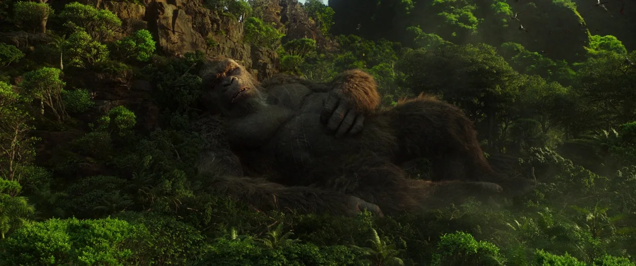 Godzilla Vs. Kong (2021) movie still: wide — A massive ape rests upon a steep, dense hillside within a lush tropical jungle. Small birds fly above…
