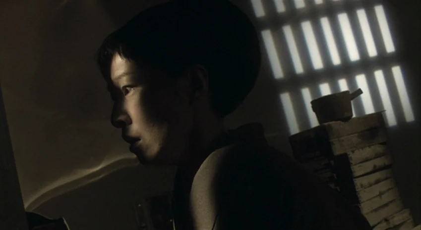 Rampo Noir (2005) movie still: closeup — A dimly lit storage area with stacked wooden crates and barred window shadows. A person stands in profile…