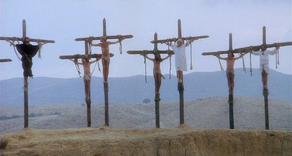 Life of Brian (1979) movie still: wide — Six wooden crosses stand in a line on a barren hilltop under cloudy skies. Multiple men are bound to the…