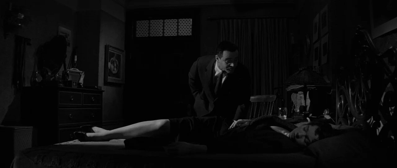 The Apartment (1960) movie still: medium — A dimly lit room with traditional furniture, a heavy wood dresser, and dark curtains. A man in a suit…