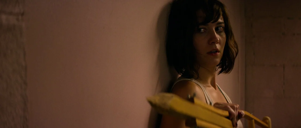 10 Cloverfield Lane (2016) movie still: medium — A dimly lit, narrow space features a concrete block wall and a smooth surface. A woman presses her…