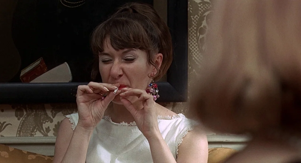 Taking Off (1971) movie still: closeup — A woman sits in a room with patterned wallpaper and a framed picture behind. She holds a cigarette between…