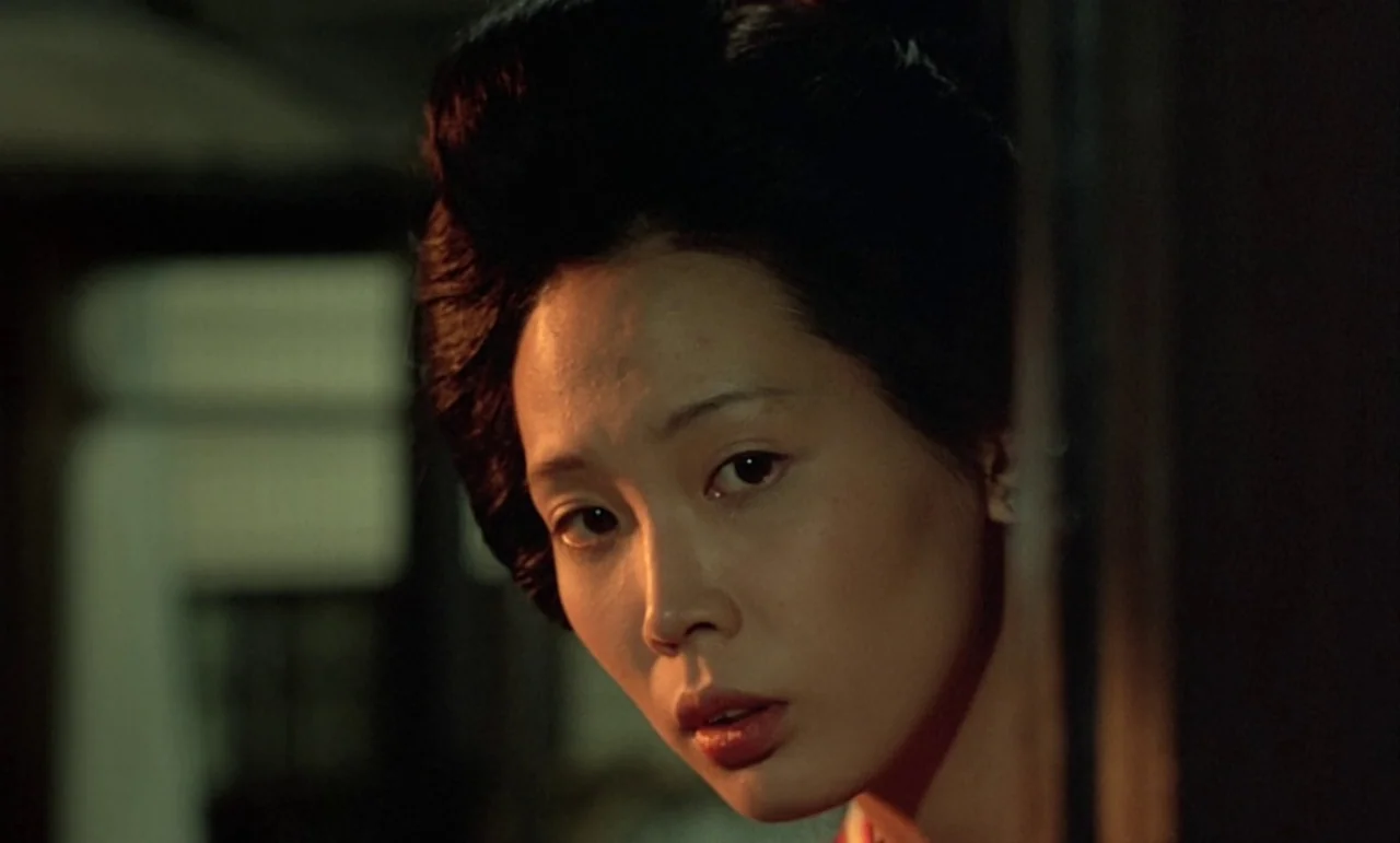 In The Realm of the Senses (1976) movie still: closeup — A dimly lit indoor space with a dark, obscured background behind a door frame. A woman with…