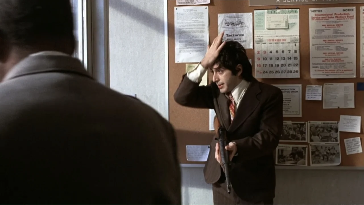 Dog Day Afternoon (1975) movie still: medium — A cluttered office wall covered with flyers and a calendar sits behind a man. A man in a suit stands…
