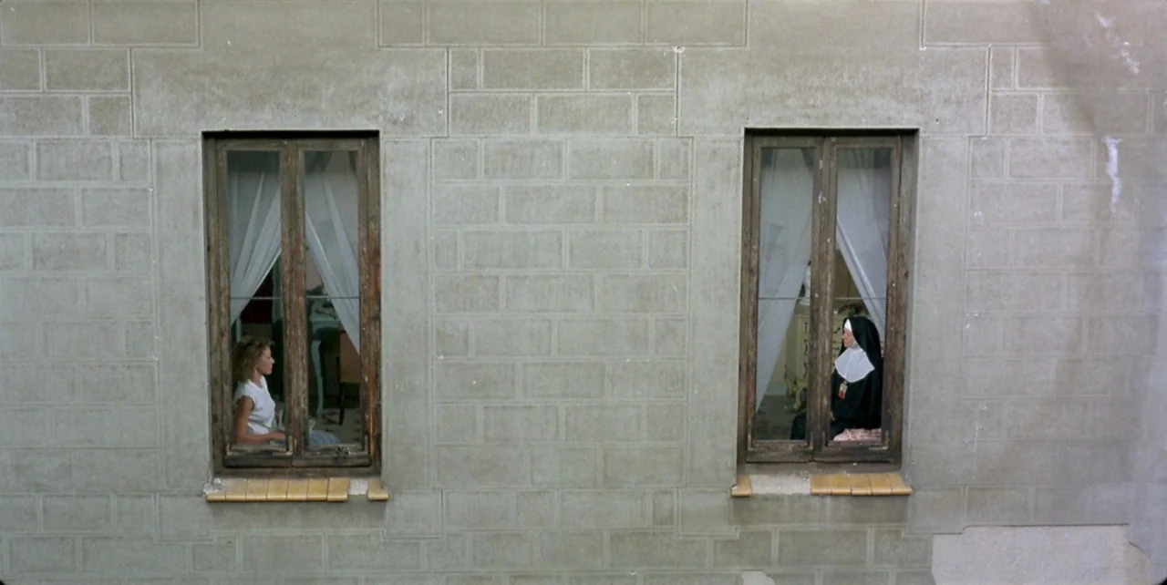 Dark Habits (1983) movie still: wide — A flat stone building wall features two symmetrical windows separated by a masonry pillar. A young woman and…