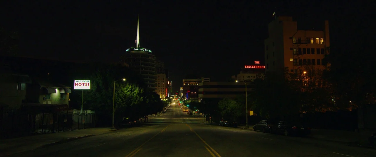 Nightcrawler (2014) movie still: establishing — An empty urban road at night stretches toward the center of the frame. Illuminated signs for the…
