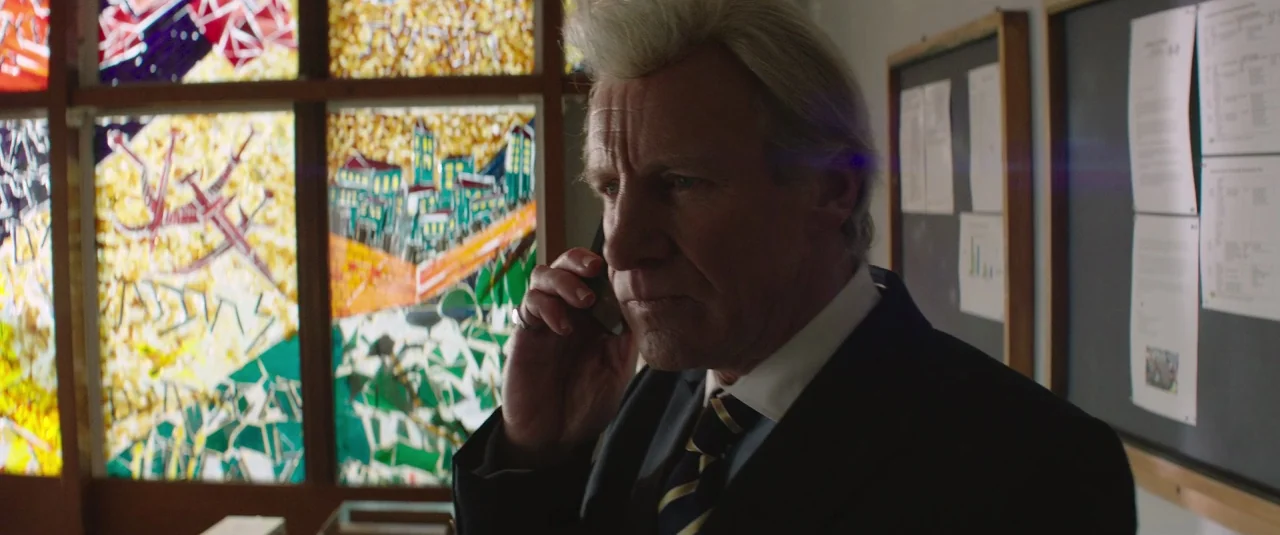 Mindhorn (2016) movie still: medium — An office interior features a wall adorned with large, multi-colored stained glass panels. An older man in a…