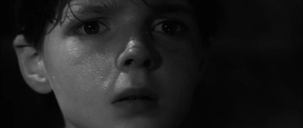 The Innocents (1961) movie still: closeup — A close-up shot of a young boy in a dark environment. His face is illuminated from the front while the…
