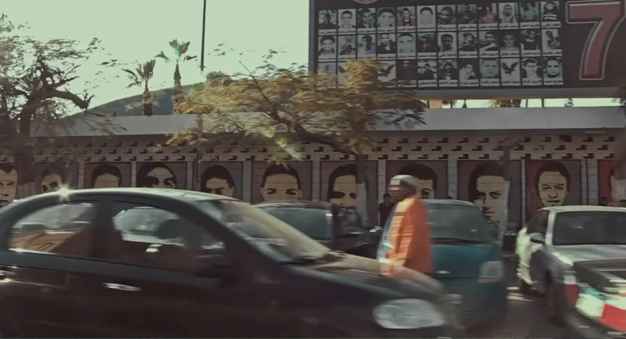 Cairo Jazzman (2017) movie still: wide — A city street features a long concrete wall decorated with many painted portrait murals. A man in an orange…