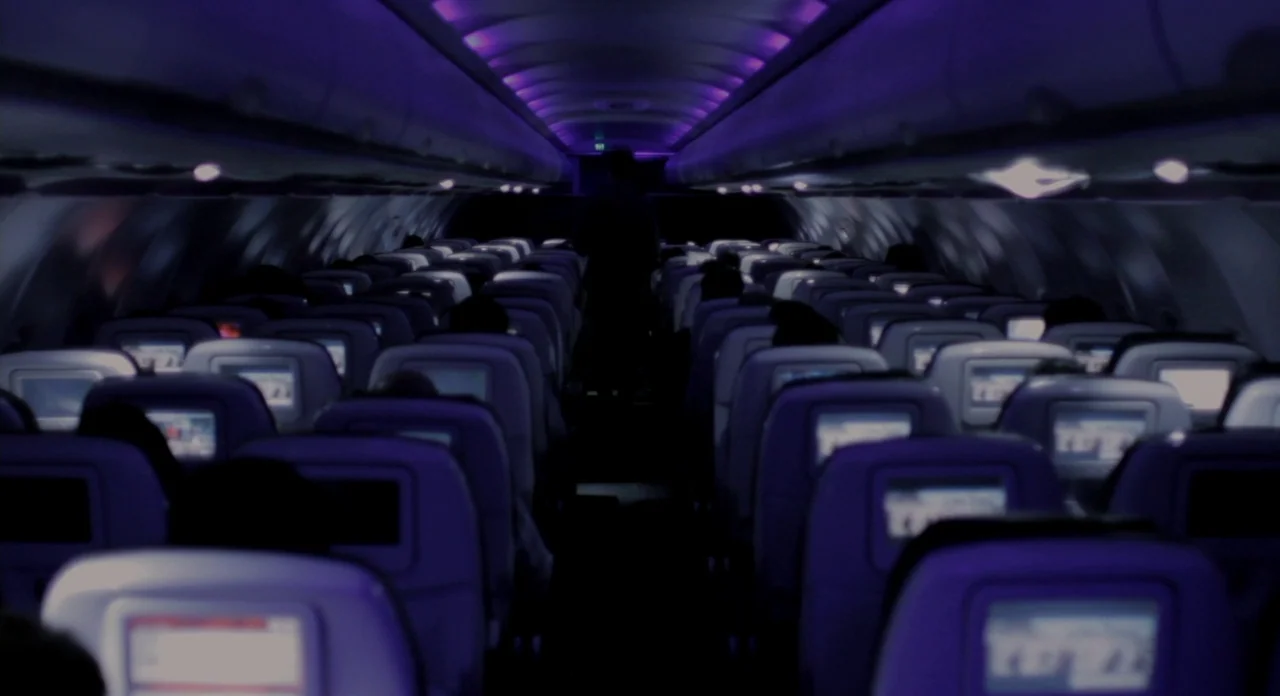 Sound Of My Voice (2011) movie still: wide — A dark airplane cabin viewed down the center aisle features rows of seats. Multiple seatback screens…
