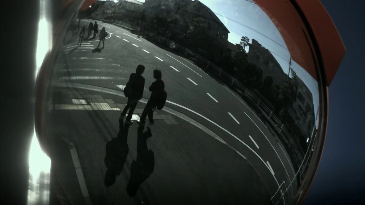 Confessions (2010) movie still: wide — A fish-eye convex mirror reflects an asphalt road intersection on a clear day. Two figures walk near each…