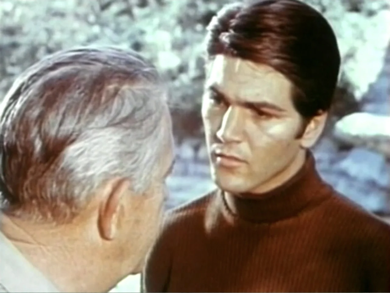 In the Year 2889 (1969) movie still: medium — An outdoor setting features blurred greenery in the background behind two male subjects. One man in a…