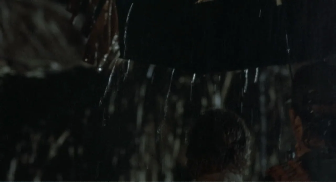 Firewalker (1986) movie still: medium — A dark outdoor space during a heavy rainstorm at night. Two people stand close together while huddled under…