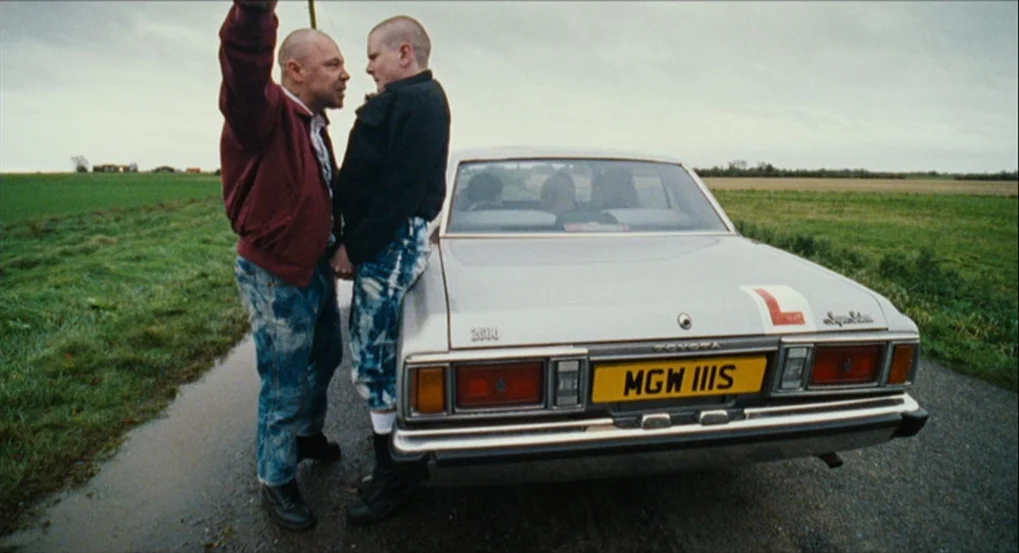 This Is England (2006) movie still: fullbody — A rural asphalt road cuts through a green field under an overcast grey sky. Two men with shaved heads…