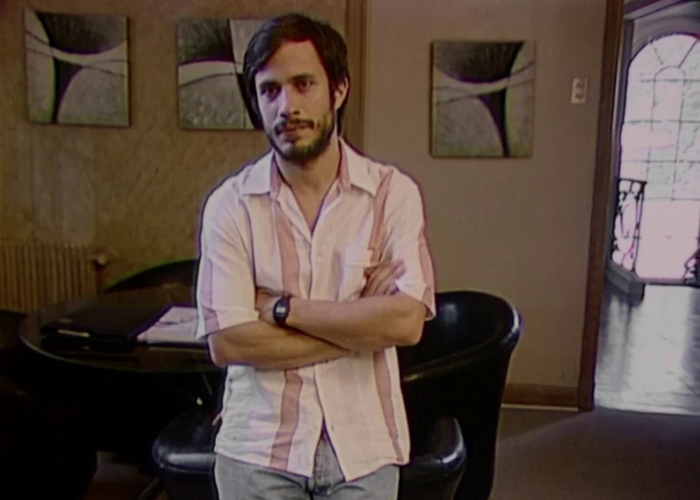 No (2012) movie still: medium — A residential living room contains modern abstract wall art and dark furniture. A bearded man in a striped shirt…