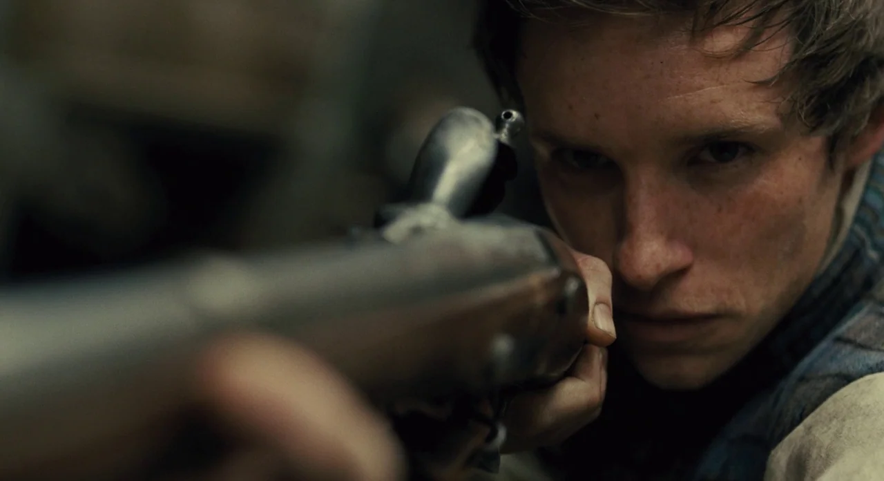 Les Misérables (2012) movie still: closeup — An outdoor setting featuring a gritty, worn barricade of wooden debris. A young man aims a flintlock…