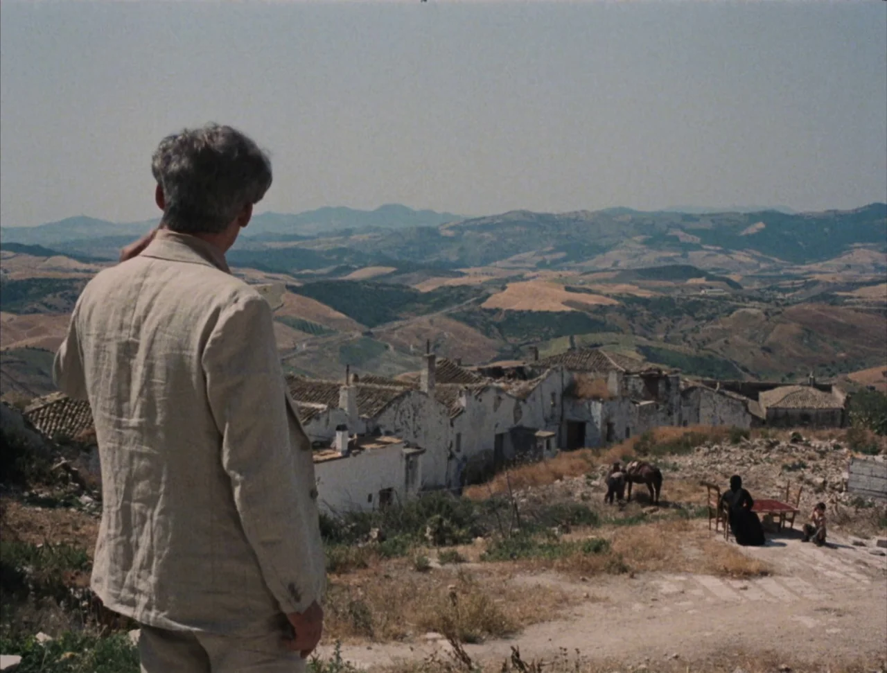 Christ Stopped at Eboli (1979) movie still: medium — A man looks over a rugged hillside toward a cluster of weathered white houses. In the distance,…