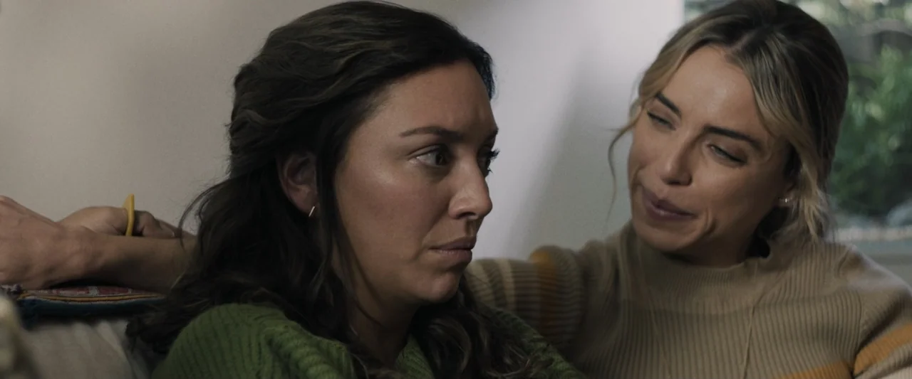 Sign Here (2017) movie still: medium — A domestic interior with a couch and blurred background textures. Two women interact closely while seated…